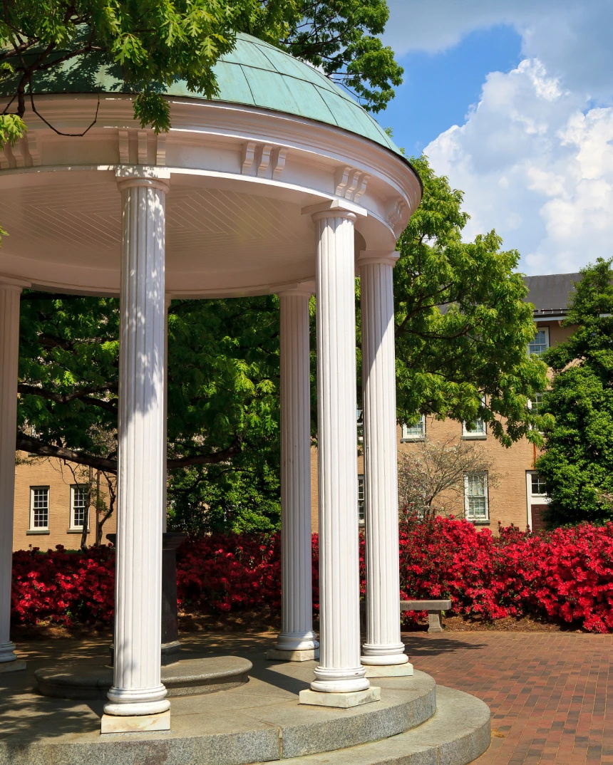White columned gazebo surrounded by red flowers and trees on a sunny day