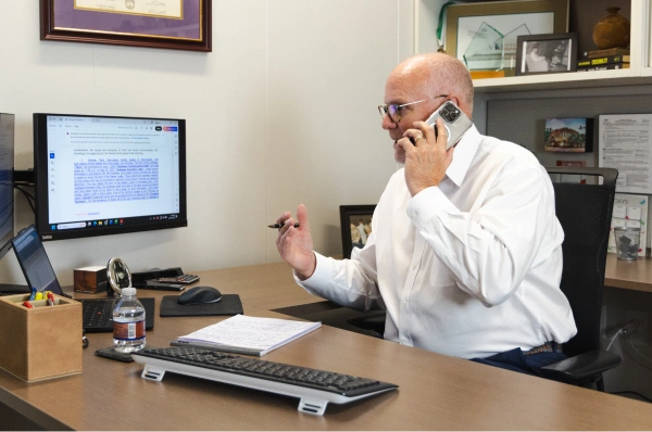 An older man sits at his desk talking on the phone while reviewing documents on his computer and taking notes