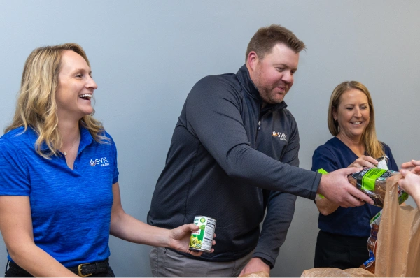 Three coworkers hand food items into a bag while smiling and helping with a group task