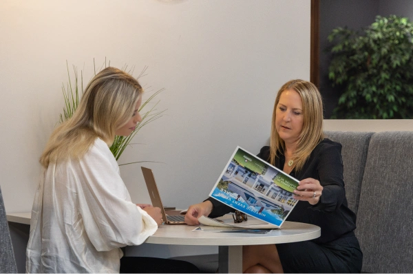 Two women sit at a small table reviewing printed materials together, with a laptop open beside them